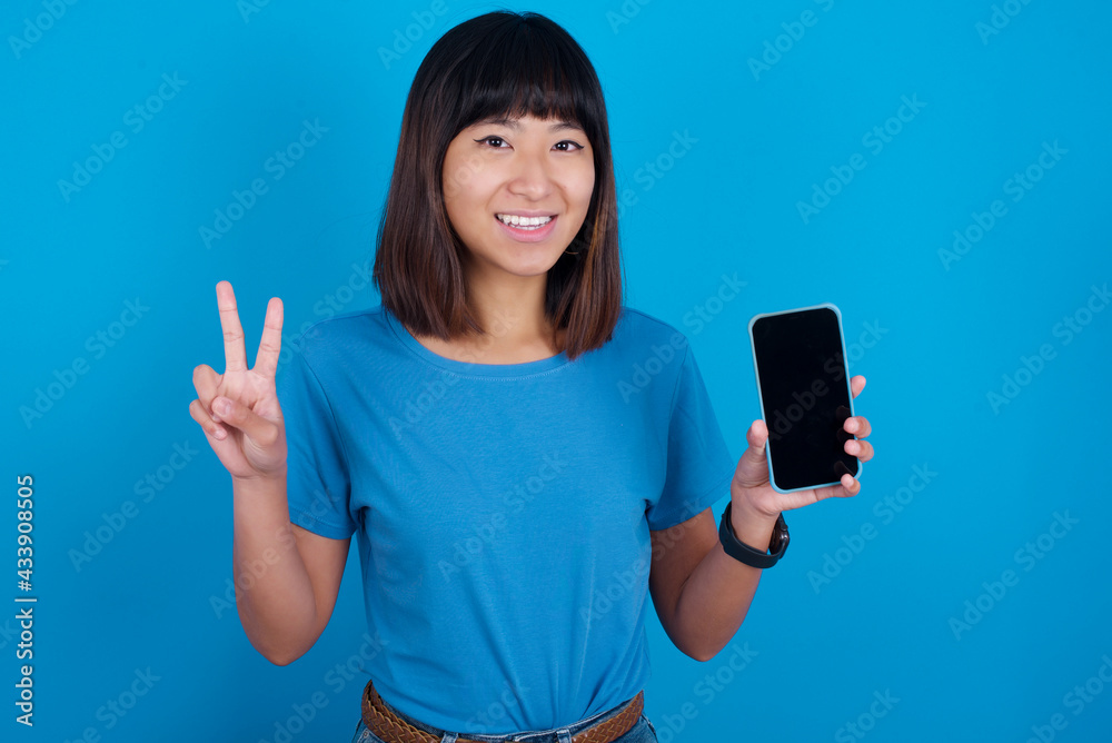 young beautiful asian girl wearing blue t-shirt against blue background holding modern device showing v-sign young beautiful asian girl wearing blue t-shirt against blue background holding modern device showing v-sign