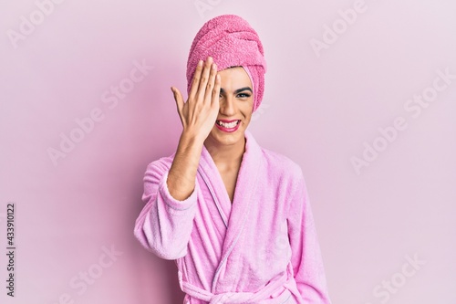 Young man wearing woman make up wearing shower towel on head and bathrobe covering one eye with hand, confident smile on face and surprise emotion. © Krakenimages.com