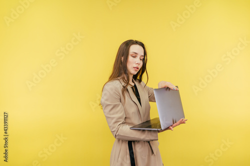 Business woman in a beige suit opens a laptop to work with a serious face on a yellow background. Attractive woman working on laptop, isolated.