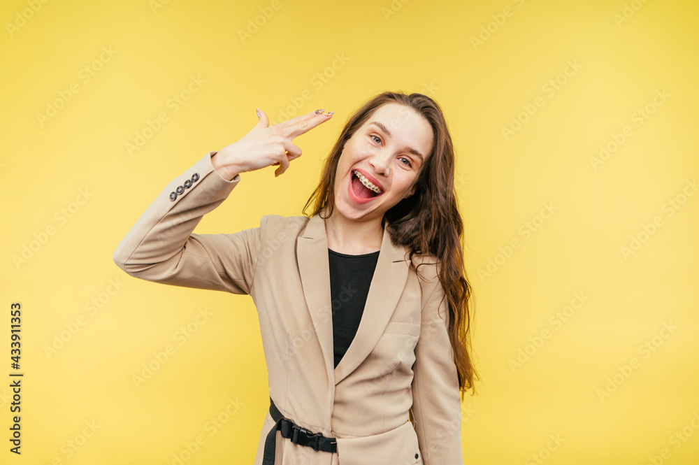 Woman in a suit stands on a beige background and shows a gesture of suicide, shoots a finger to the head, looks at the camera with a crazy smiling face. Woman in a suit stands on a beige background and shows a gesture of suicide, shoots a finger to the head, looks at the camera with a crazy smiling face.