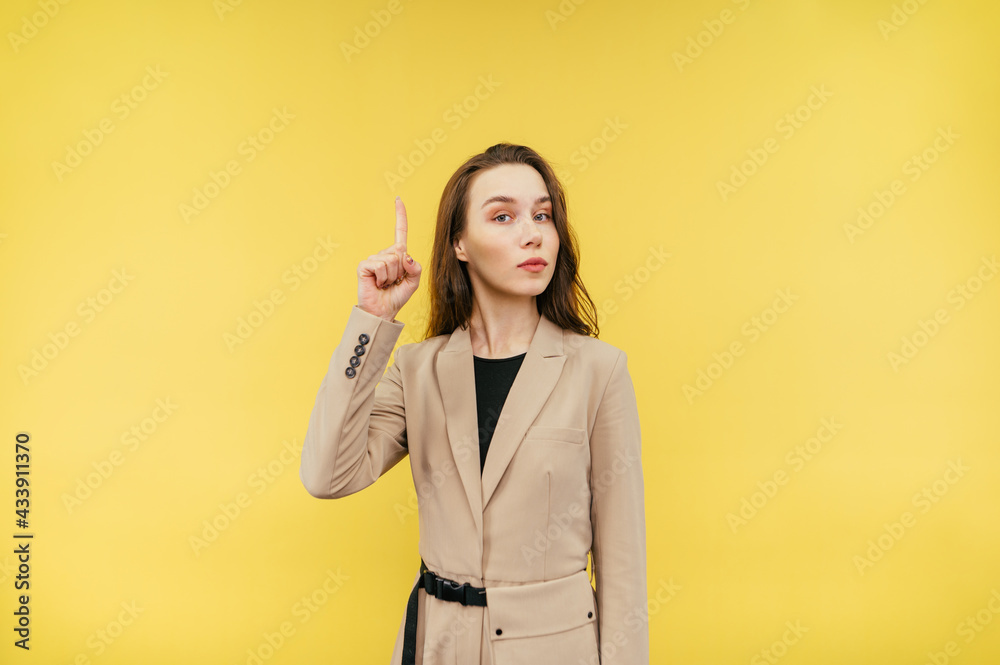 Serious woman in smart casual clothes stands on a yellow background, looks at the camera with a serious face and points a finger at copy space Serious woman in smart casual clothes stands on a yellow background, looks at the camera with a serious face and points a finger at copy space