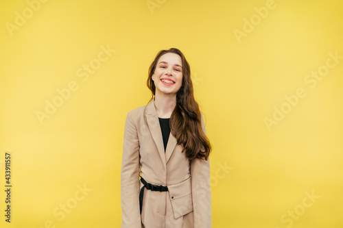Portrait of a joyful woman with braces on her teeth emotionally smiling on a yellow background and looking at the camera, wearing a beige jacket