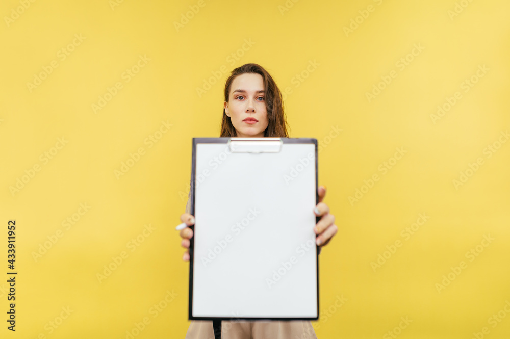 Beautiful female office worker holds a paper tablet with a white sheet and shows to the camera isolated on a yellow background. Beautiful female office worker holds a paper tablet with a white sheet and shows to the camera isolated on a yellow background.