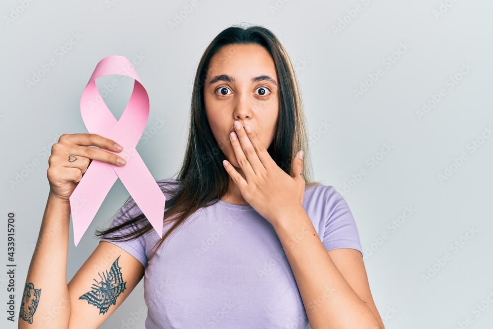 Young hispanic woman holding pink cancer ribbon covering mouth with hand, shocked and afraid for mistake. surprised expression Young hispanic woman holding pink cancer ribbon covering mouth with hand, shocked and afraid for mistake. surprised expression