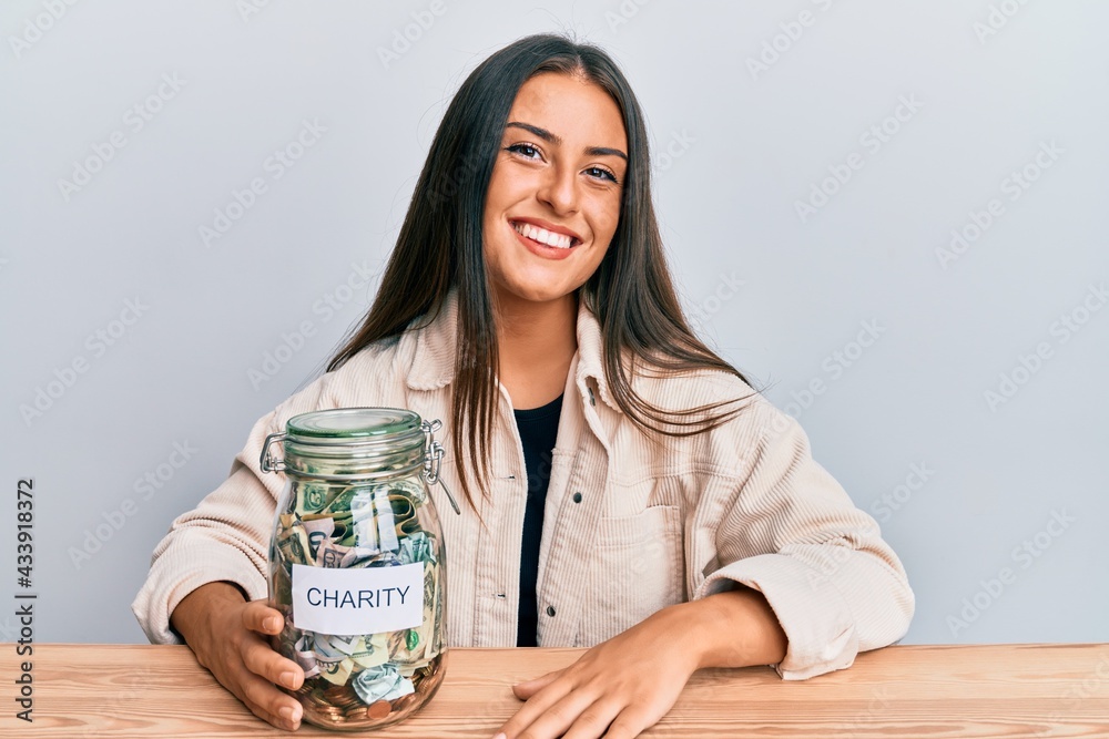 Beautiful hispanic woman holding charity jar with money looking positive and happy standing and smiling with a confident smile showing teeth Beautiful hispanic woman holding charity jar with money looking positive and happy standing and smiling with a confident smile showing teeth