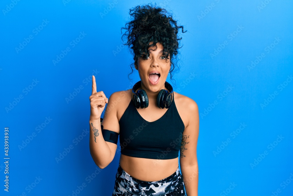 Young hispanic woman wearing gym clothes and using headphones pointing finger up with successful idea. exited and happy. number one. Young hispanic woman wearing gym clothes and using headphones pointing finger up with successful idea. exited and happy. number one.