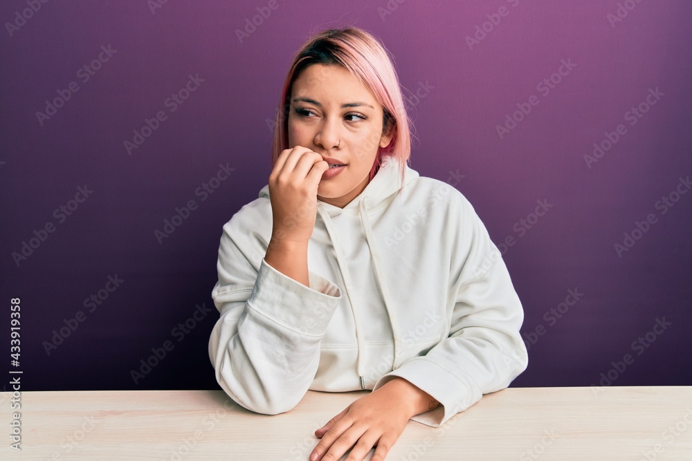 Hispanic woman with pink hair wearing casual sweatshirt sitting on the table looking stressed and nervous with hands on mouth biting nails. anxiety problem. Hispanic woman with pink hair wearing casual sweatshirt sitting on the table looking stressed and nervous with hands on mouth biting nails. anxiety problem.
