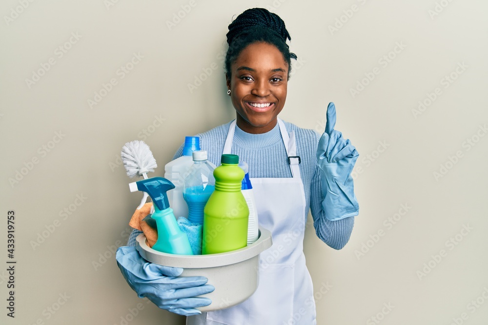 African american woman with braided hair wearing apron holding cleaning products surprised with an idea or question pointing finger with happy face, number one