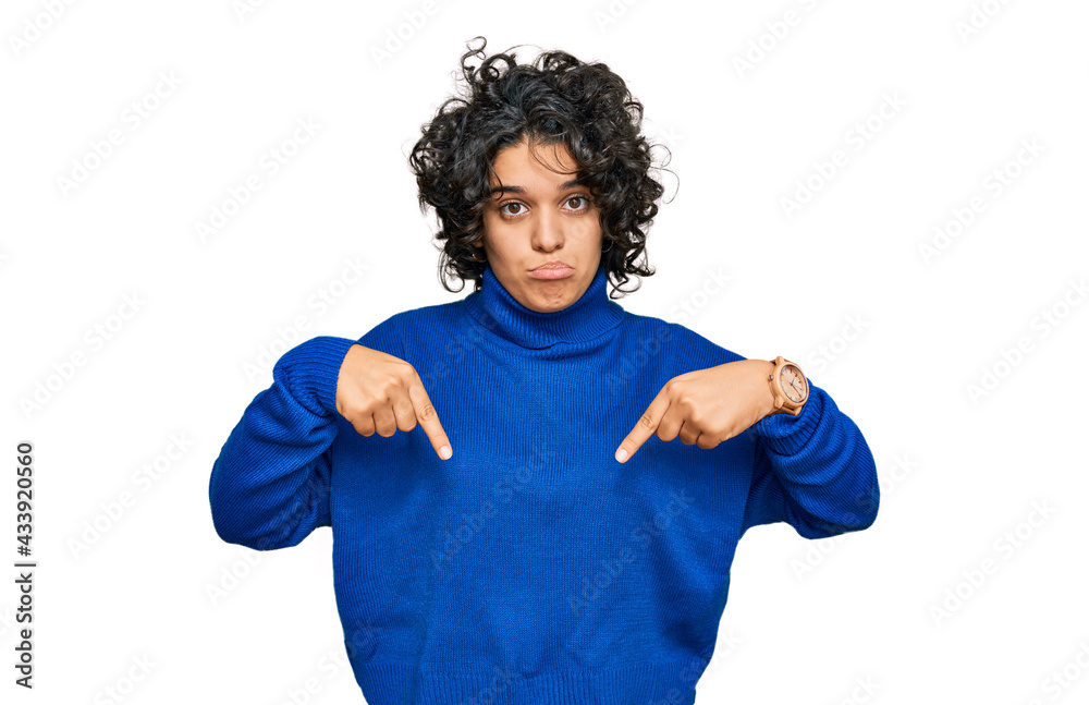 Young hispanic woman with curly hair wearing turtleneck sweater pointing down looking sad and upset, indicating direction with fingers, unhappy and depressed. Young hispanic woman with curly hair wearing turtleneck sweater pointing down looking sad and upset, indicating direction with fingers, unhappy and depressed.