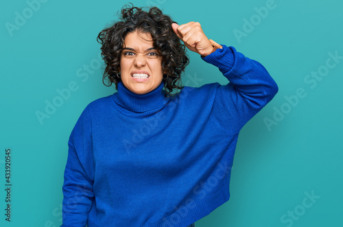 Young hispanic woman with curly hair wearing turtleneck sweater angry and mad raising fist frustrated and furious while shouting with anger. rage and aggressive concept. © Krakenimages.com