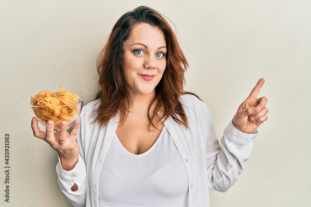 Young caucasian woman holding bowl of uncooked pasta smiling happy pointing with hand and finger to the side Young caucasian woman holding bowl of uncooked pasta smiling happy pointing with hand and finger to the side