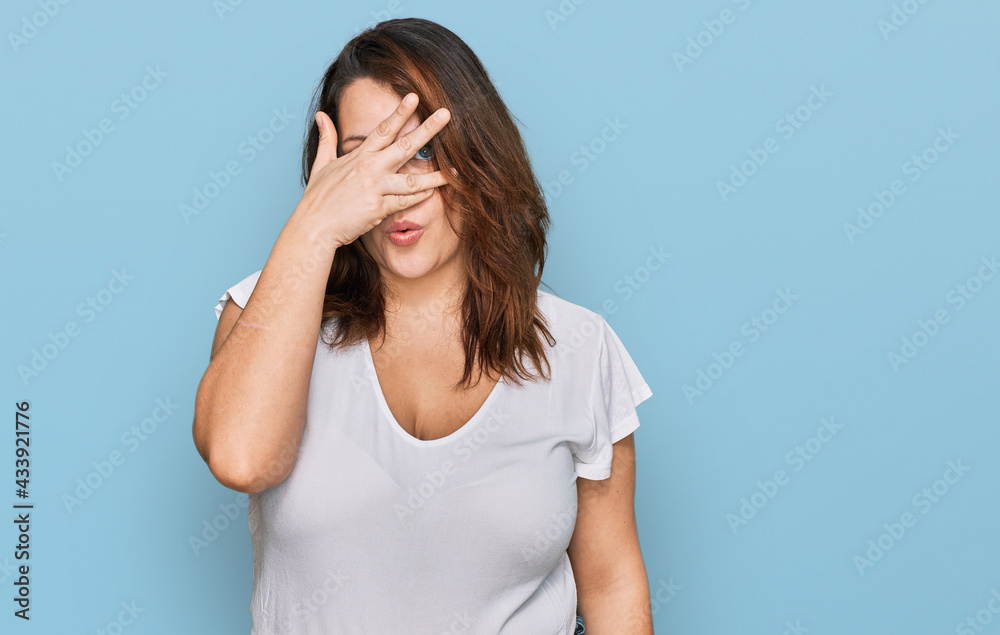 Young plus size woman wearing casual white t shirt peeking in shock covering face and eyes with hand, looking through fingers with embarrassed expression. Young plus size woman wearing casual white t shirt peeking in shock covering face and eyes with hand, looking through fingers with embarrassed expression.