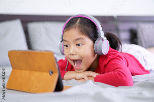 Asian child or kid girl learn from home or people study online on computer tablet and wearing headphone to e-learning communication by pad with open mouthed wow and sleep lie on stomach on bed in room