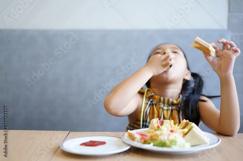 Asian child hungry or kid girl smile to enjoy happy eating bread or sandwich with ketchup and vegetable salad on dish and table at school or home kitchen and restaurant for breakfast food on arm focus photo