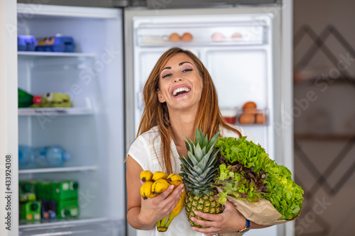 Young woman with fruits and vegetables beside fridge in kitchen. purchase box full of vegetables standing beside fridge