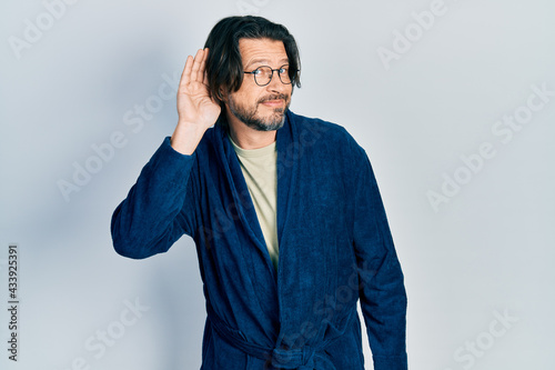 Middle age caucasian man wearing bathrobe and glasses smiling with hand over ear listening an hearing to rumor or gossip. deafness concept.