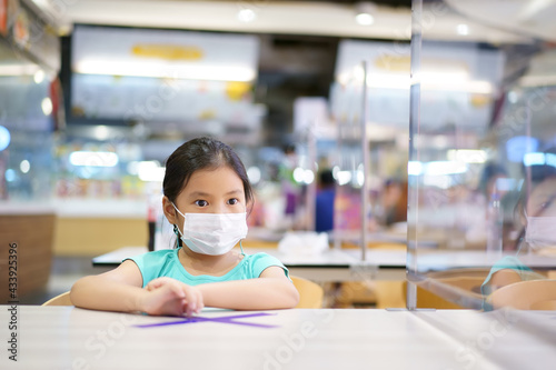 Asian child or kid girl wearing white face mask sitting with protection glass partition and cross mark on table for protect coronavirus covid-19 to social distancing at food court or school restaurant