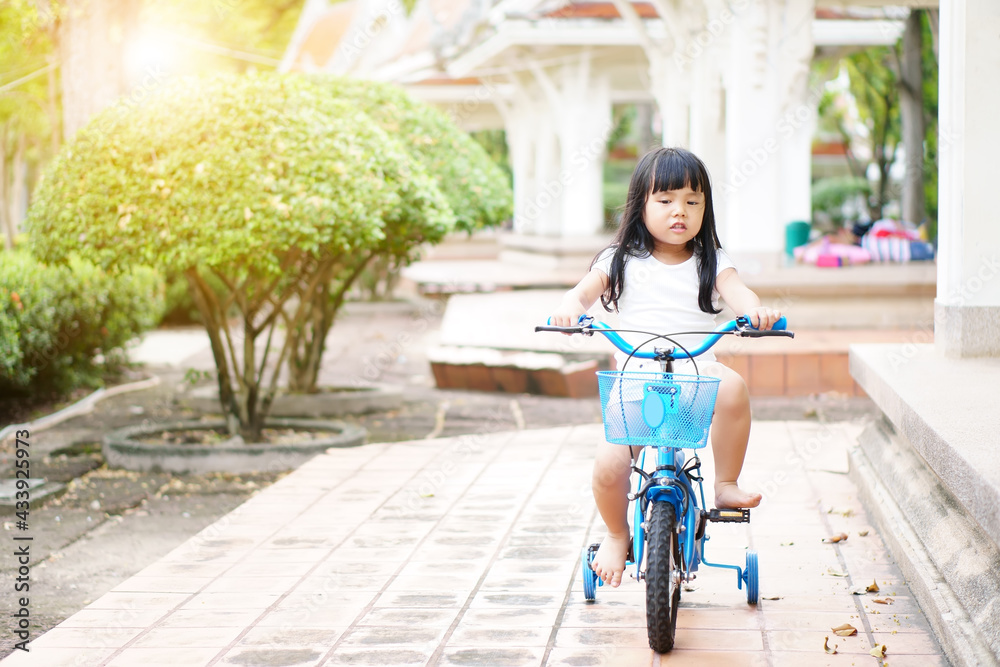 Asian child preschool or kid girl wearing white shirt and cycling on blue bike or riding bicycle and happy on public park with green garden and tree to enjoy sport exercise with fun on summer holiday