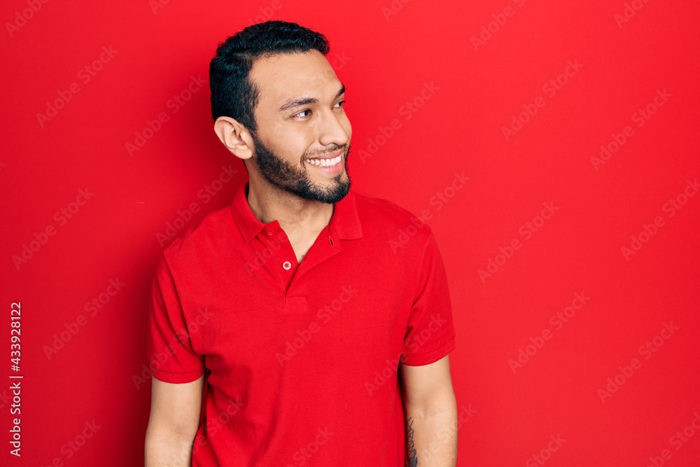 Hispanic man with beard wearing casual red t shirt looking away to side with smile on face, natural expression. laughing confident. Hispanic man with beard wearing casual red t shirt looking away to side with smile on face, natural expression. laughing confident.
