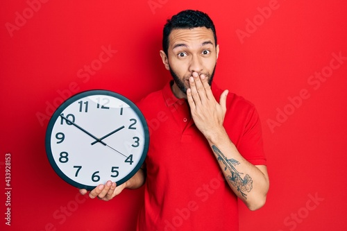 Hispanic man with beard holding big clock covering mouth with hand, shocked and afraid for mistake. surprised expression
