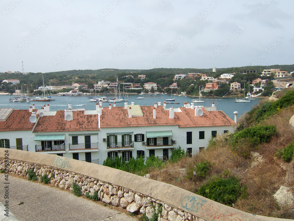 View from a hill above residential houses to the small harbor of Menorca with many sailboats and fishing boats lying in the water of the ocean. In the background you can see beach houses on a hill