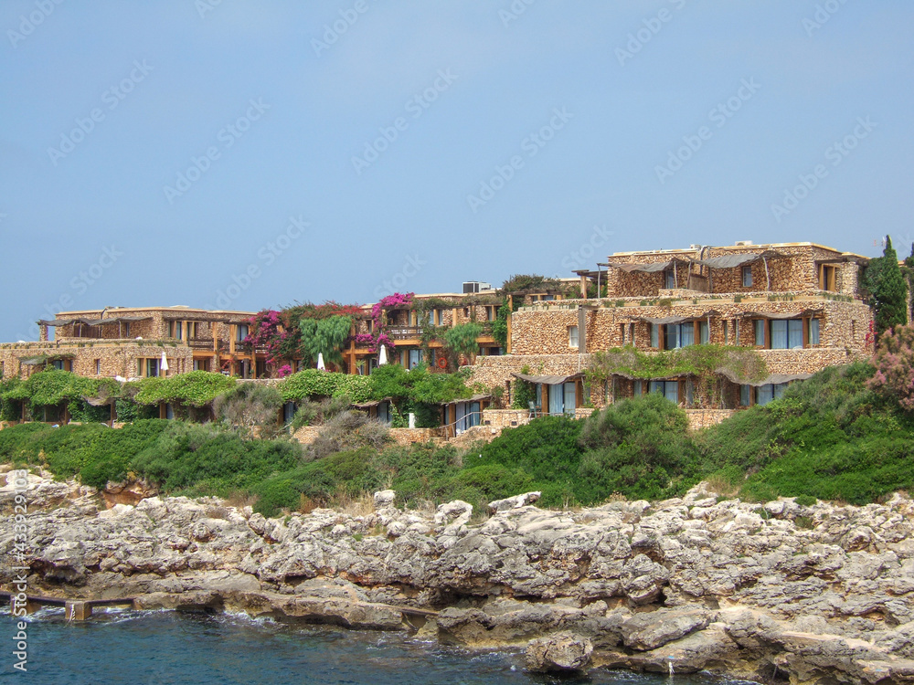 Beach houses built of orange sandstone on the rocky coast of Menorca. Above the cliff to the sea and small planted hills stand some luxury villas overlooking the sea