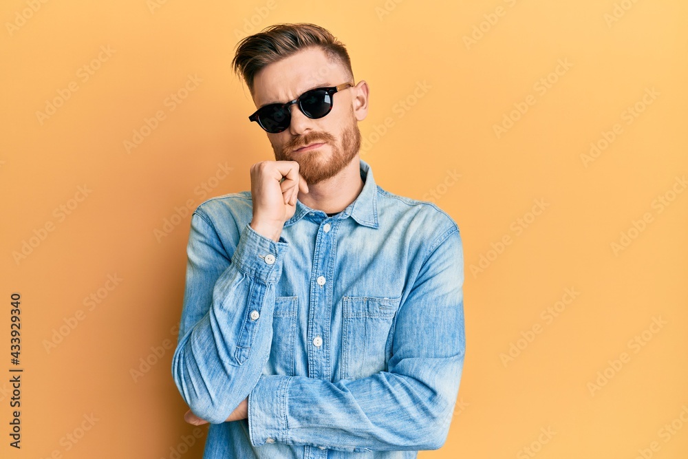 Young redhead man wearing stylish sunglasses serious face thinking about question with hand on chin, thoughtful about confusing idea Young redhead man wearing stylish sunglasses serious face thinking about question with hand on chin, thoughtful about confusing idea