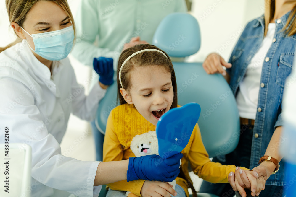 Girl looking in the mirror after dental procedure while mother standing near her for support. Girl looking in the mirror after dental procedure while mother standing near her for support.
