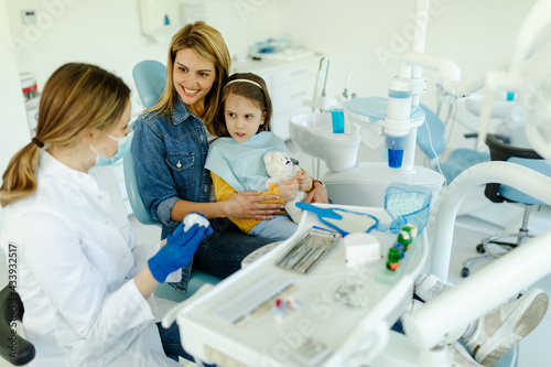 Little cute smiling girl is sitting in dental chair with mother in clinic, office.
