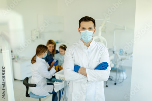 Male dentist, standing in dental office, wearing mask, white coat and gloves while posing.