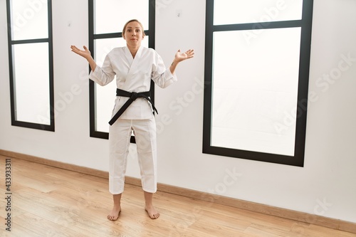 Caucasian young blonde woman wearing karate kimono and black belt clueless and confused expression with arms and hands raised. doubt concept.