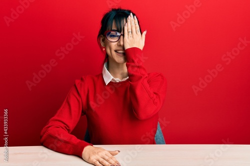 Young brunette woman with bangs wearing glasses sitting on the table covering one eye with hand, confident smile on face and surprise emotion.