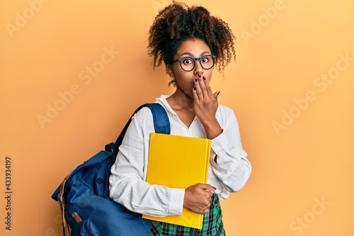Beautiful african american woman with afro hair wearing school bag and holding books covering mouth with hand, shocked and afraid for mistake. surprised expression