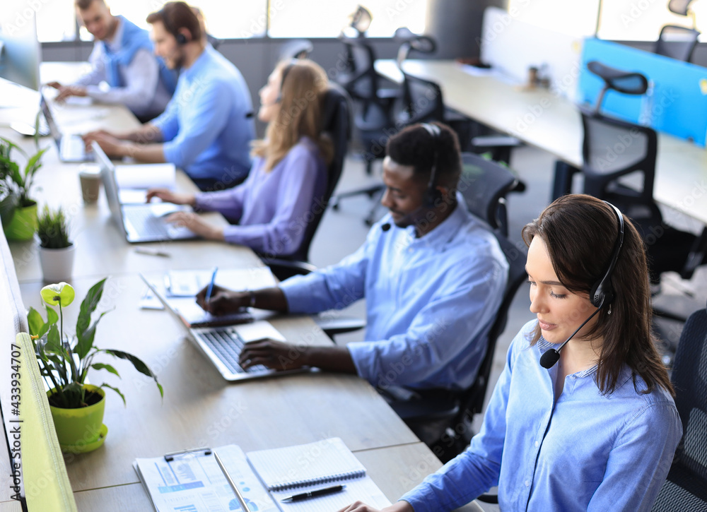 Female customer support operator with headset and smiling, with collegues at background. Female customer support operator with headset and smiling, with collegues at background.