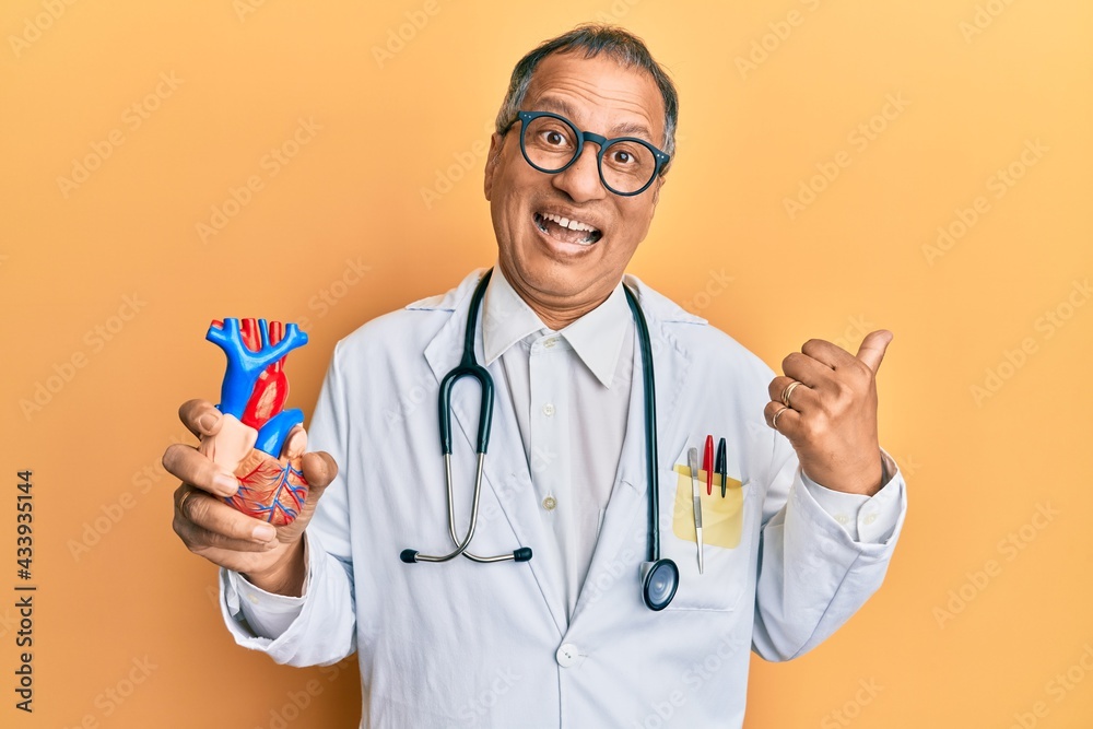 Middle age indian man wearing doctor uniform holding heart pointing thumb up to the side smiling happy with open mouth Middle age indian man wearing doctor uniform holding heart pointing thumb up to the side smiling happy with open mouth