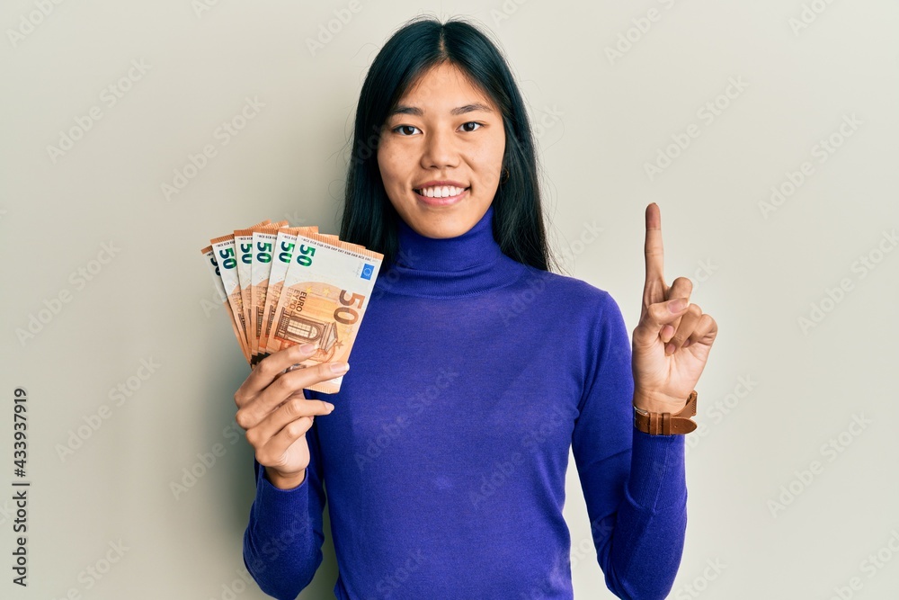 Young asian woman holding bunch of 50 euro banknotes smiling with an idea or question pointing finger with happy face, number one Young asian woman holding bunch of 50 euro banknotes smiling with an idea or question pointing finger with happy face, number one
