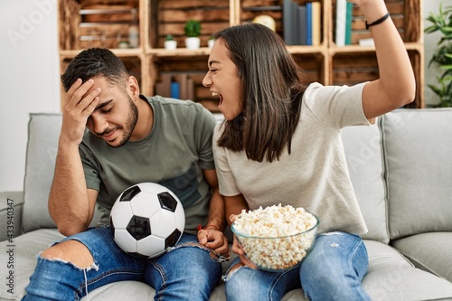 Young latin couple watching soccer match eating porpcorn at home.