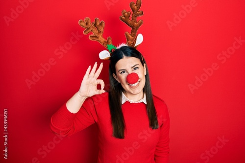 Young hispanic woman wearing deer christmas hat and red nose smiling positive doing ok sign with hand and fingers. successful expression.