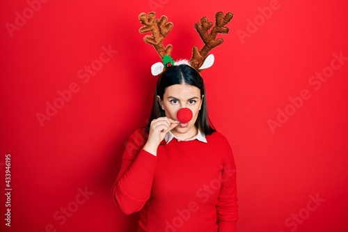 Young hispanic woman wearing deer christmas hat and red nose mouth and lips shut as zip with fingers. secret and silent, taboo talking