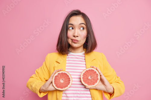 Horizontal shot of thoughtful Asian woman holds two grapefruit halves concentrated away wears stylish clothes isolated over pink background. Natural nutrition and juicy citrus fruit concept. photo