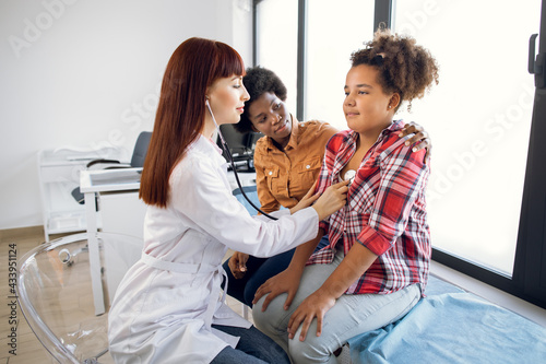 Flu, cough, asthma concept. Female pediatrician doctor listens to lungs of African-American 12-aged girl with stethoscope. Sick teen mixed race girl with asthma, supporting by her worried mom photo