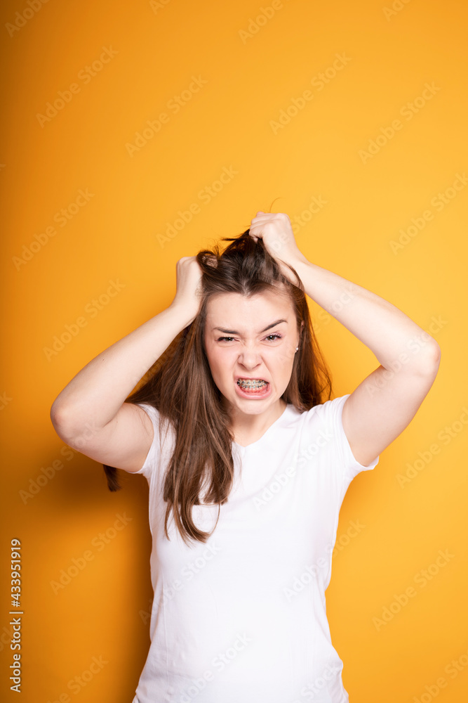 Angry young girl with braces pulls out the hair on her head from pain in the teeth and in the head while standing on a yellow background Angry young girl with braces pulls out the hair on her head from pain in the teeth and in the head while standing on a yellow background