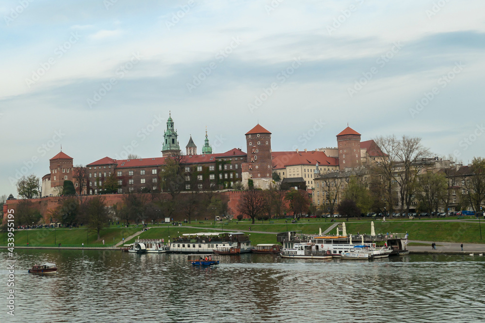 A panoramic view on the iconic Wawel Castle in Cracow, Poland and the Vistula river flowing under the castle. A few birds flying above the river. A few boats docked on the rivers bend. City tour. A panoramic view on the iconic Wawel Castle in Cracow, Poland and the Vistula river flowing under the castle. A few birds flying above the river. A few boats docked on the rivers bend. City tour.