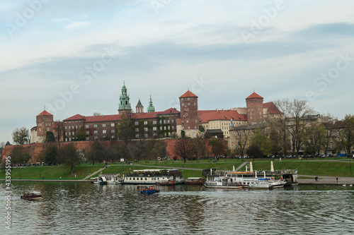 A panoramic view on the iconic Wawel Castle in Cracow, Poland and the Vistula river flowing under the castle. A few birds flying above the river. A few boats docked on the rivers bend. City tour.