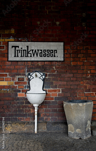 Public white drink water fountain on a red brick wall. The German script painted above reads 