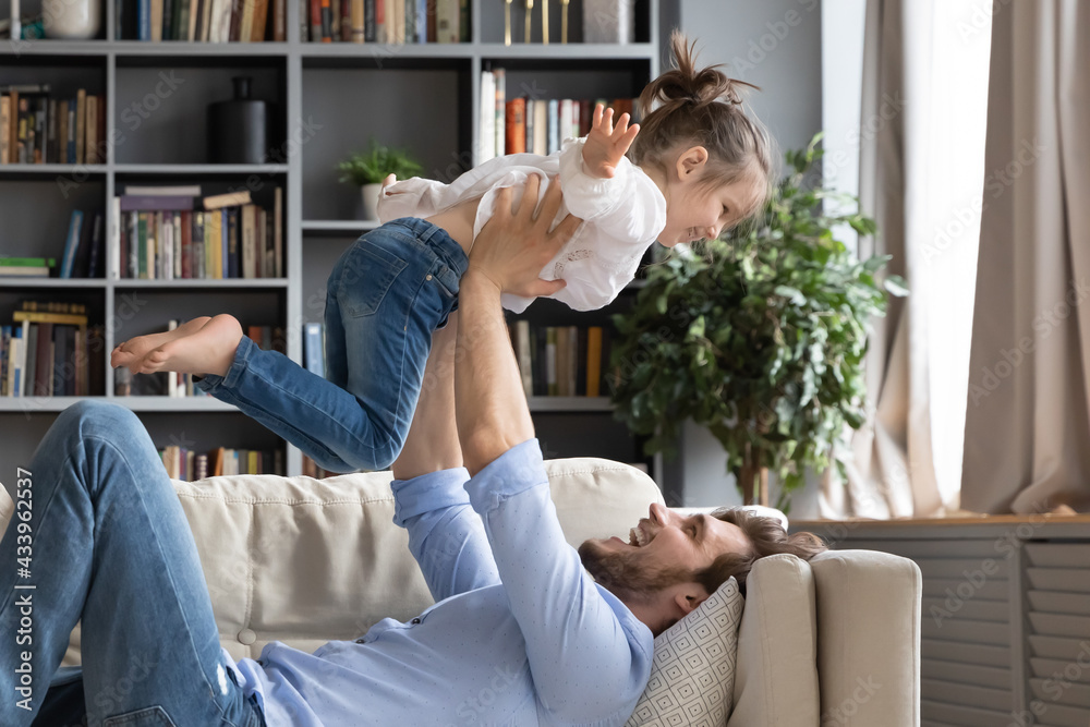 Happy young father lying on couch at home lift up in air imitate plane fly with little daughter. Smiling dad play with small excited girl child, relax together in living room, feel playful in game.