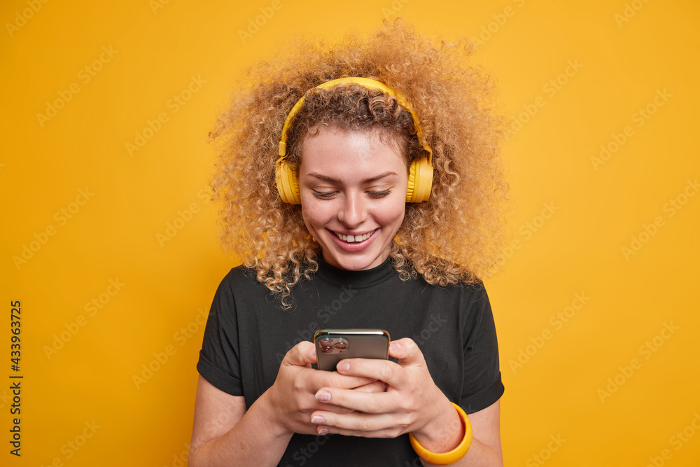Best music app for free. Cheerful curly haired woman in casual black t shirt uses modern wireless headphones and smartphone has access to popular songs enjoys playlist isolated over yellow wall Best music app for free. Cheerful curly haired woman in casual black t shirt uses modern wireless headphones and smartphone has access to popular songs enjoys playlist isolated over yellow wall