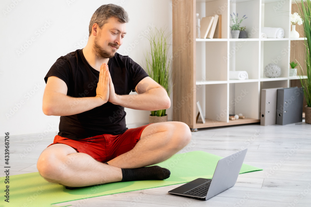 Fototapeta premium Yoga practice. Meditating man. Online training. Video conference. Peaceful smiling guy in casual sportswear sitting lotus pose holding namaste hands looking laptop light home interior.