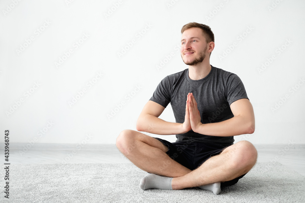Balance yoga. Happy man. Home meditation. Peaceful mind. Smiling guy in t-shirt shorts sitting lotus pose namaste hands on floor light wall interior background copy space. Balance yoga. Happy man. Home meditation. Peaceful mind. Smiling guy in t-shirt shorts sitting lotus pose namaste hands on floor light wall interior background copy space.