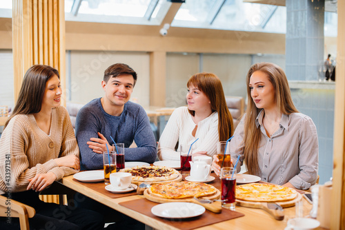 A group of young cheerful friends is sitting in a cafe talking and eating pizza. Lunch at the pizzeria.